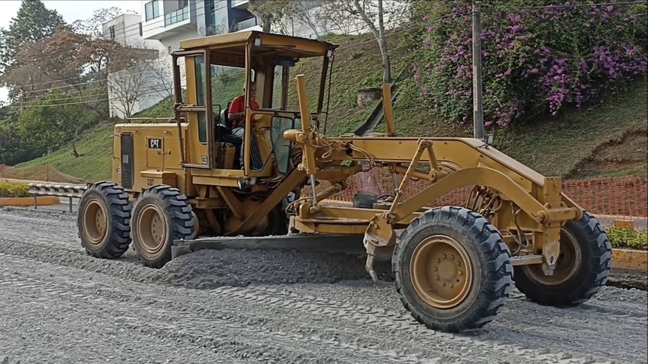 Motoniveladora Caterpillar 12G esparciendo grava y compactadora compactando grava en pavimentación