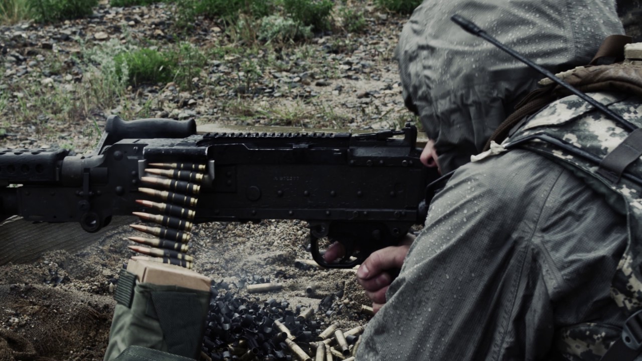 Shot of a soldier firing a belt-fed machine gun from a sitting position.