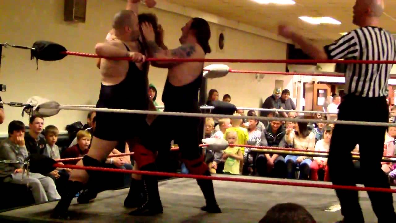 husky wrestler rubs his opponent's face in his tag-partner's armpit