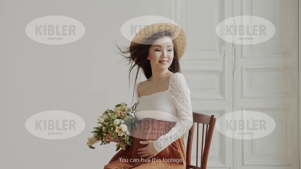 Pregnant asian woman sits on chair and poses with flowers indoors in daytime