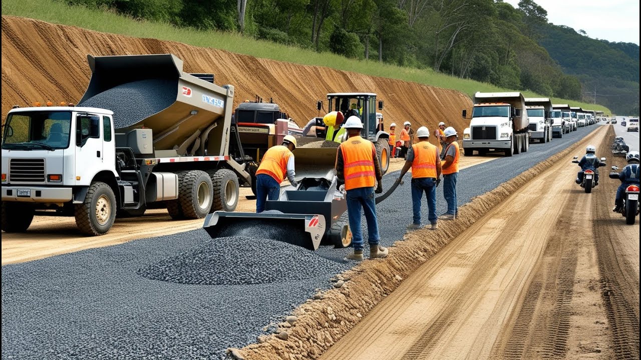 Construction machinery actively paving a road. A dump truck is unloading gravel into a gravel paver