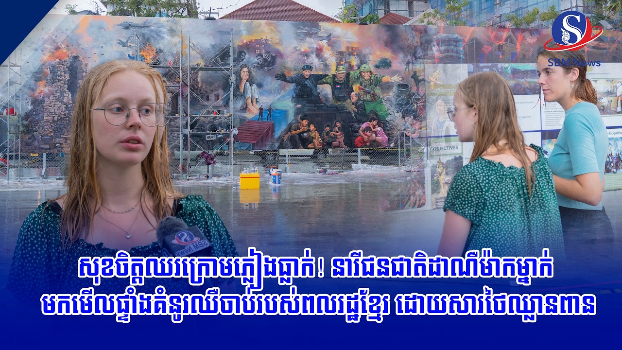 សុខចិត្តឈរក្រោមភ្លៀងធ្លាក់! នារីជនជាតិដាណឺម៉ាកម្នាក់ មកមើលផ្ទាំងគំនូរឈឺចាប់របស់ពលរដ្ឋខ្មែរ ដោយសារថៃឈ
