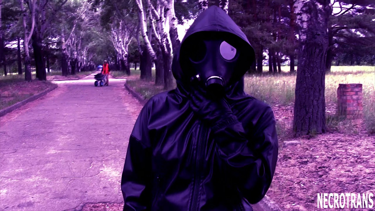 A girl in a black leather suit and an Israeli gas mask walks in a park near the river bank.