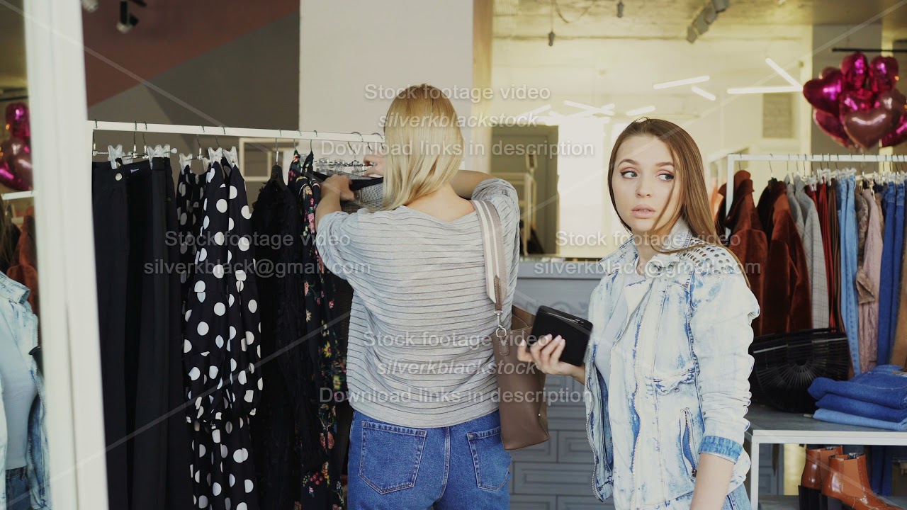 Young woman stealing wallet from female customer choosing clothes in shop. Thief looking around