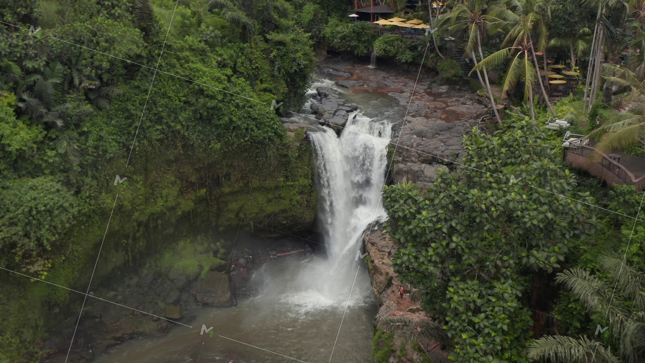 Aerial shot flying towards a large waterfall in the Bali jungle with outdoors restaurant on the
