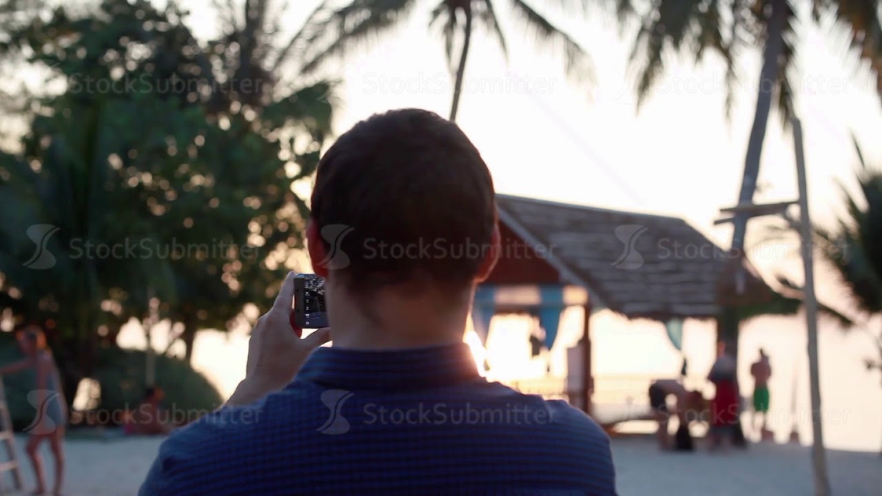 Young handsome man standing on beach taking photo with smart phone of sunset over Koh Phangan