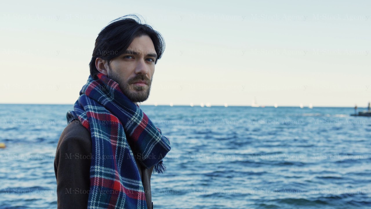 Portrait shot of a melancholic good-looking man in glasses and motly scarf looking at the sea and