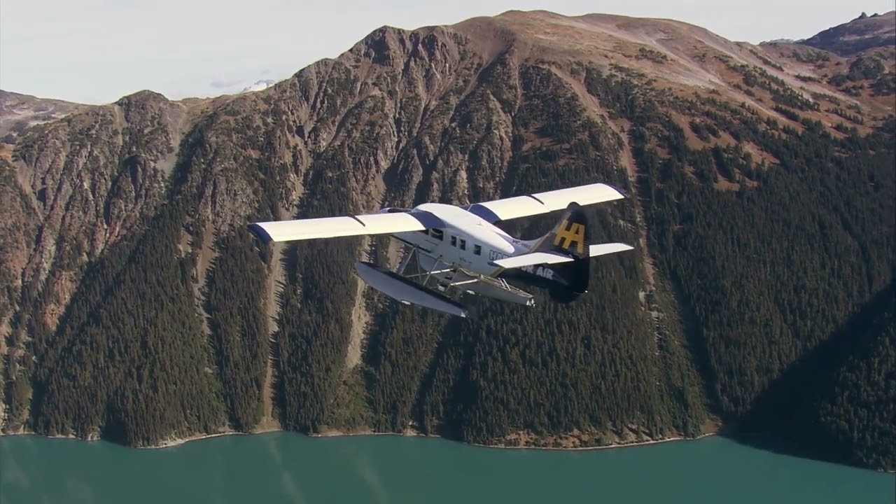 Aerial shot of a Harbour Air seaplane flying over a blue lake towards forest covered
