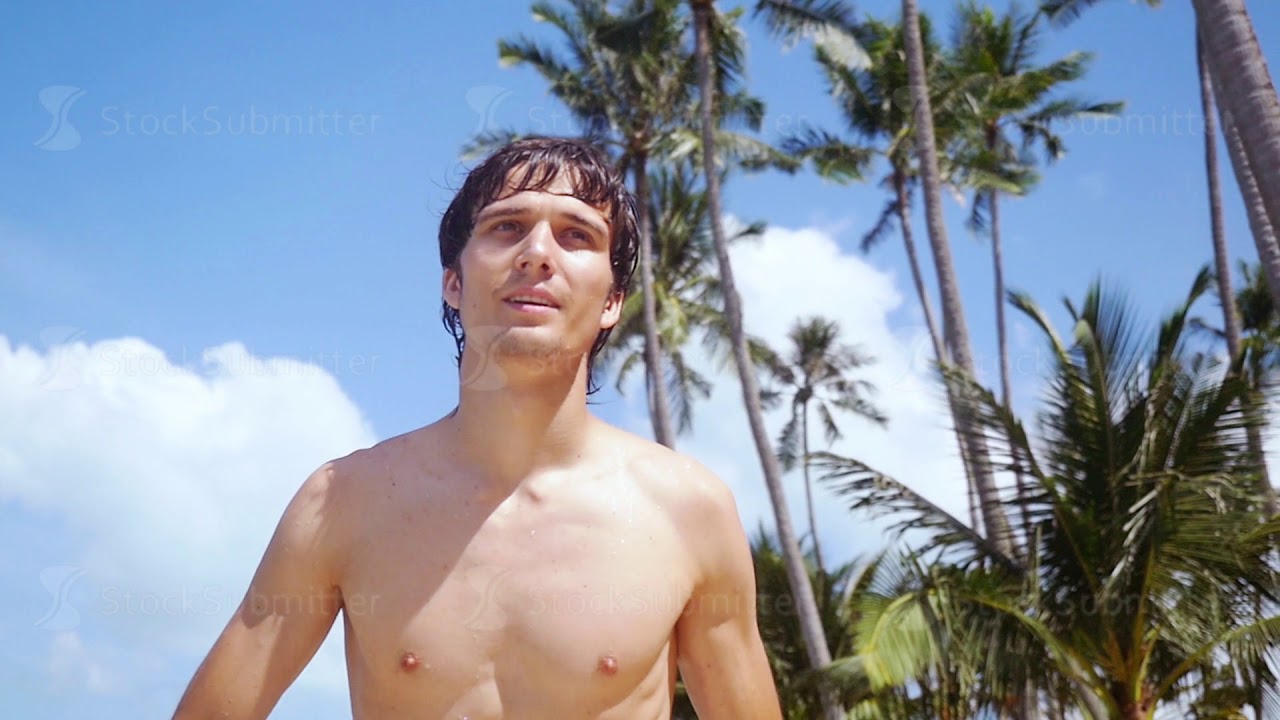 Young sexy handsome man running on the beach at sunny day against palm trees and the blue sky on the