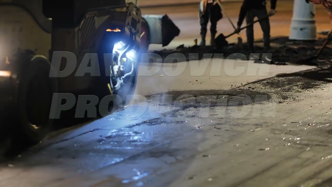 Road roller compacting hot asphalt on city street at night, workers repairing