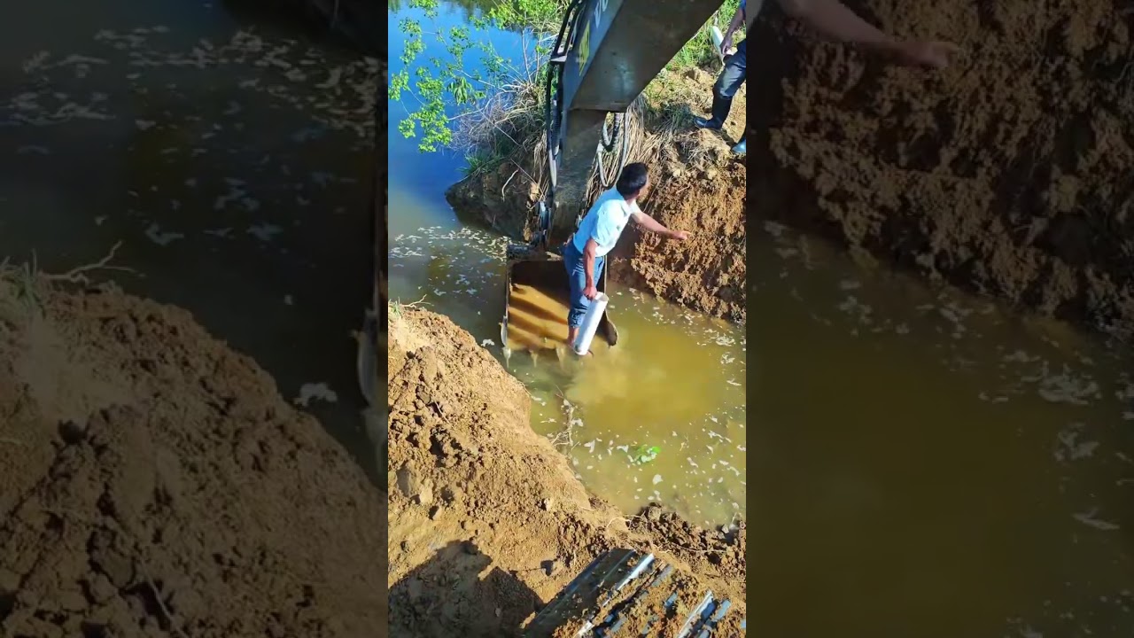 A man on a backhoe bucket pulls a plastic pipe out of the water