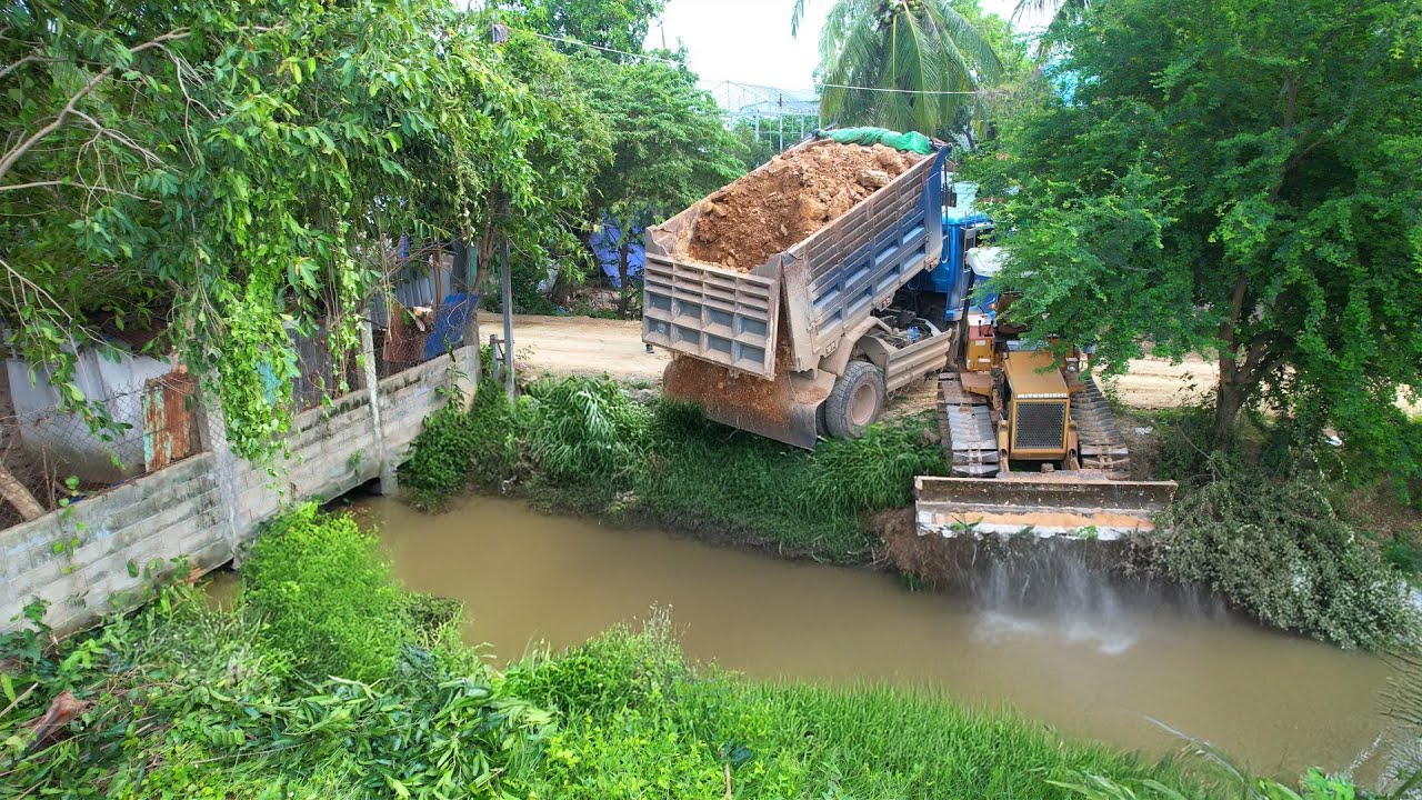 Powerful Machinery at Work: Dirt Truck Empties Load, Bulldozer Masterfully Manages Riverbank Fill
